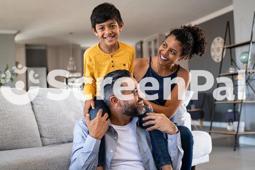 Cheerful African Mother And Indian Father Playing With Son At Home Cute Boy Enjoying Sitting On Father Shoulder While Looking At Camera Middle Eastern Family Having Fun Together On The Sofa At Home