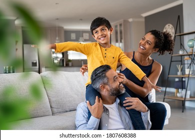 Cheerful Indian Son Sitting On Father Shoulder Playing At Home With African Mother Playful Little Boy Enjoying Spending Time With Parents At Home Flying Child Enjoying Playing With His Ethnic Family