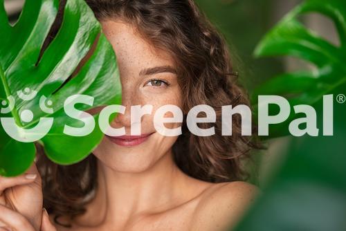 Close Up Face Of Beautiful Young Woman Covering Her Face By Green Monstera Leaf While Looking At Camera Portrait Of Beauty Woman With Natural Makeup And Freckles Standing Behind Big Green Leaves