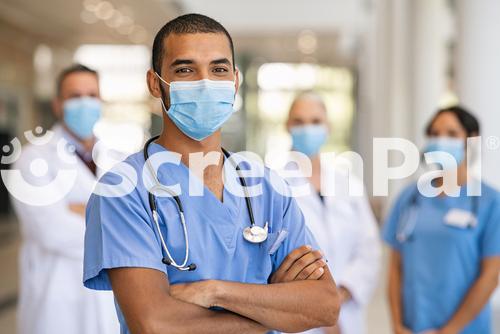 Confident Multiethnic Male Nurse In Front Of His Medical Team Looking At Camera Wearing Face Mask During Covid 19 Outbreak Happy And Proud Indian Young Surgeon Standing In Front Of His Colleagues