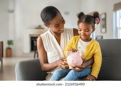 Smiling Mature African American Mother Helping Daughter Sitting On Lap Putting Money In Piggy Bank Cute Little Black Girl Saving Money By Adding A Coin In Piggy Bank With Mother At Home 