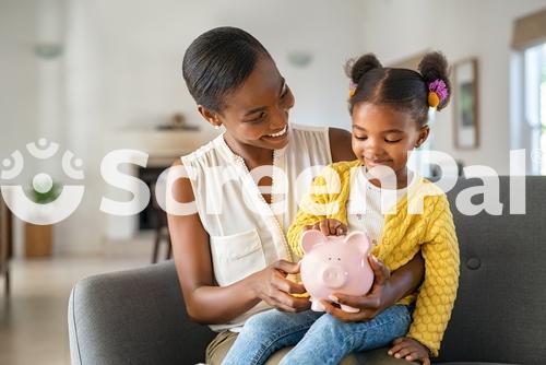 Smiling Mature African American Mother Helping Daughter Sitting On Lap Putting Money In Piggy Bank Cute Little Black Girl Saving Money By Adding A Coin In Piggy Bank With Mother At Home 