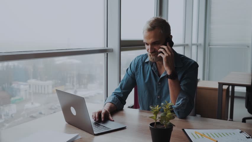 Male Businessman Is Discussing A Handheld Document On A Mobile Phone With A Pleased Smile While Seated At His Desk In The Office Working On The Computer