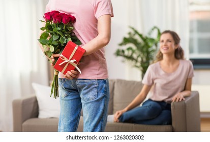 Couple Relationships And People Concept   Happy Woman Looking At Man Hiding Flowers And Gift Behind His Back At Home