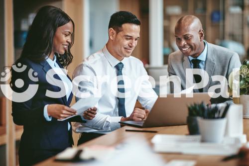 Smiling Group Of Diverse Businesspeople Going Over Paperwork Together And Working On A Laptop At A Table In An Office