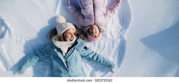 Top View Portrait Of Cheerful Mother With Small Daughter Lying In Snow In Winter Nature